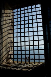 Ein verrostetes Gitter einer Gefängniszelle, im Hintergrund gehen das blaue Meer und der blaue Himmel ineinander über.
