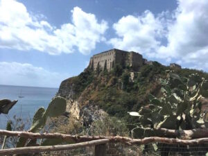 Sicht auf eine Burg, hochoben auf einem Felsen. Davor das Meer, im Hintergrund leicht bewölkter, blauer Himmel.